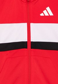 Röd zip-up jacka med vita och svarta horisontella ränder över bröstet. Har strukturerad tyg och en vit logotyp i övre vänstra hörnet.