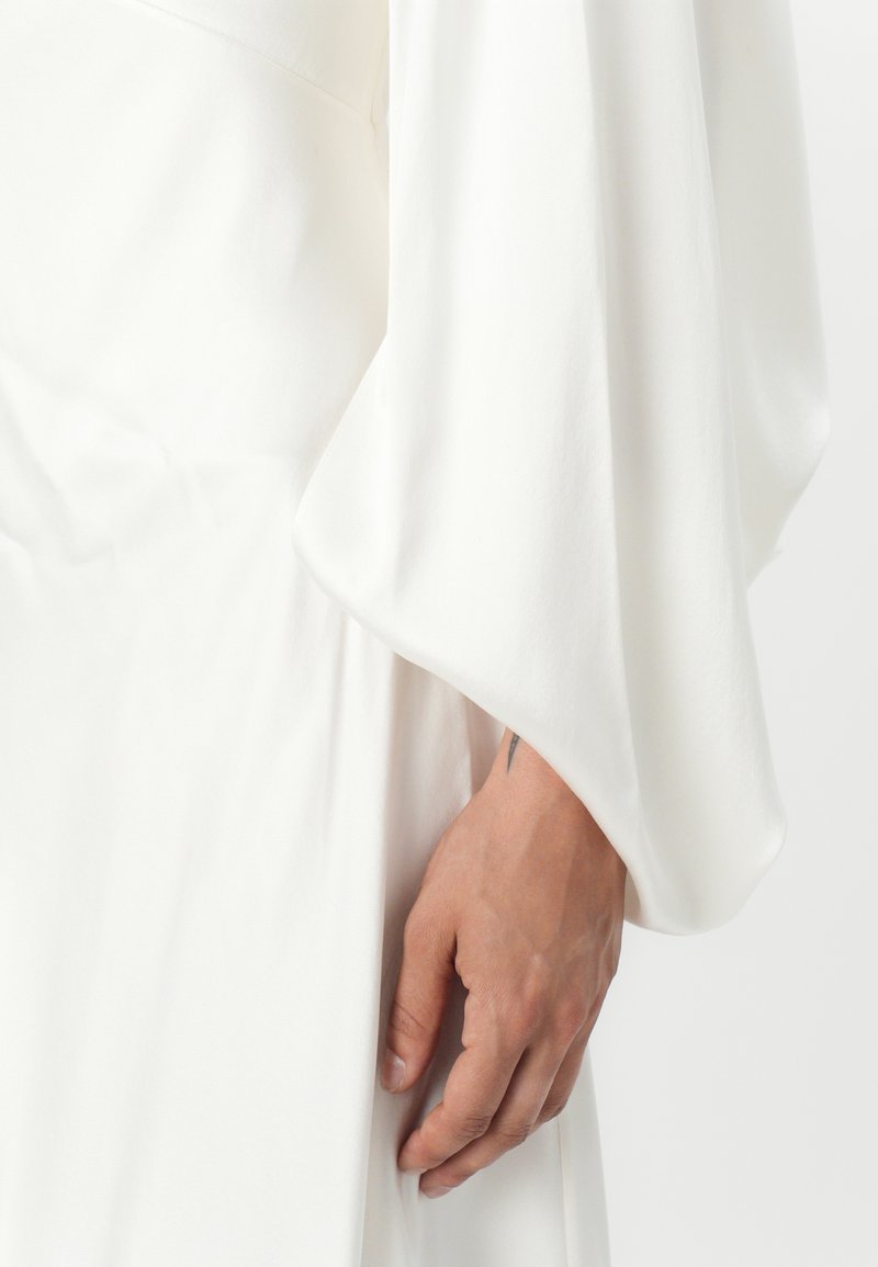 Robe blanche avec de longues manches fluides et une texture douce. Les bords des manches sont lisses ; la main repose près de la taille. Le teint de la peau est visible près du tissu.