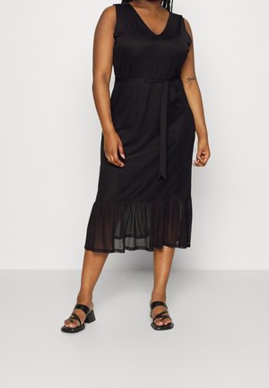 Schwarzes Midi-Kleid mit V-Ausschnitt, ärmellosem Design, Taillenzug und durchscheinendem, rüschengesäumtem Saum. Getragen mit schwarzen High Heels.