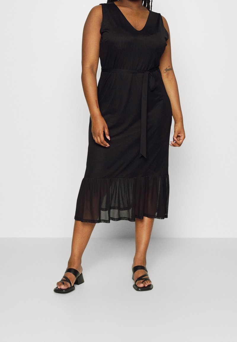 Zwart midi-jurk met een V-hals, mouwloos ontwerp, tailleband en doorschijnende gerimpelde zoom. Gemaakt in combinatie met zwarte hoge haksandalen.