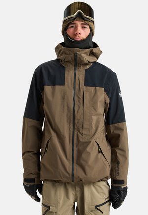 Veste imperméable marron et noire avec un col haut, une fermeture éclair à l'avant et des poches latérales. Portée avec des lunettes de ski et des gants pour les sports d'hiver.