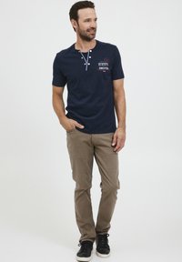 Jūrų laivynui priklausanti trumpomis rankovėmis henley marškinėliai su dviem mygtukais, šviesiai mėlyna apykaklė ir logotipo detalė. Derinami su smėlio spalvos siauromis kelnėmis ir juodais batais.