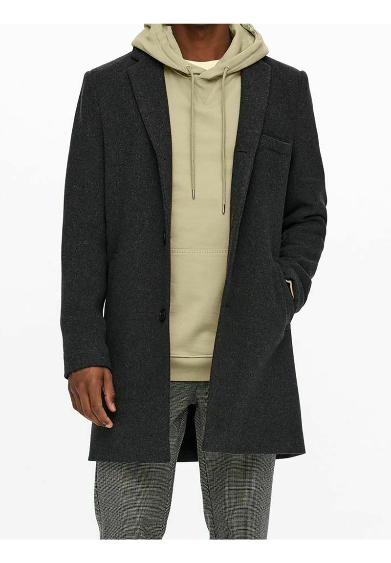 Donkergrijze wollen overjas met een effen ontwerp en twee voorkantzakken, gedragen over een lichtgroene hoodie en patroonbroeken.