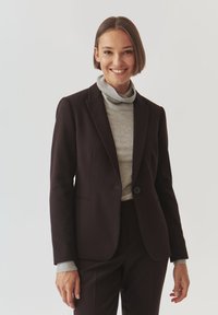 Dark brown tailored blazer with a smooth texture, single button closure, and two front pockets, worn over a light gray turtleneck.