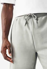 Pantalon de survêtement gris clair avec une taille à cordon de serrage, présentant une texture lisse et des poches latérales, partiellement visible avec une main près du tissu.