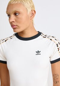 Adidas t-skjorte i hvit med svarte detaljer. Har leopardmønstrede striper på skuldrene og en liten logo på brystet. Korte ermer; tettsittende design.