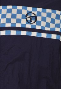Tessuto blu navy con una striscia a scacchi blu e bianca che attraversa il petto, caratterizzato da un logo circolare ricamato al centro.