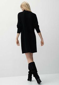 Femme aux longs cheveux blonds portant une robe noire à manches longues et des bottes à talons hauts noires et décontractées, debout devant un fond clair uni.