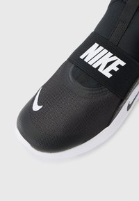 Chaussure de sport noire avec une tige en tissu texturé, semelle en caoutchouc, logo Nike blanc bien visible sur la sangle et détail de capuchon lisse à l'avant.