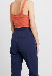 Top corto arancione ruggine con spalline spesse, abbinato a pantaloni navy a vita alta con dettaglio della tasca posteriore.