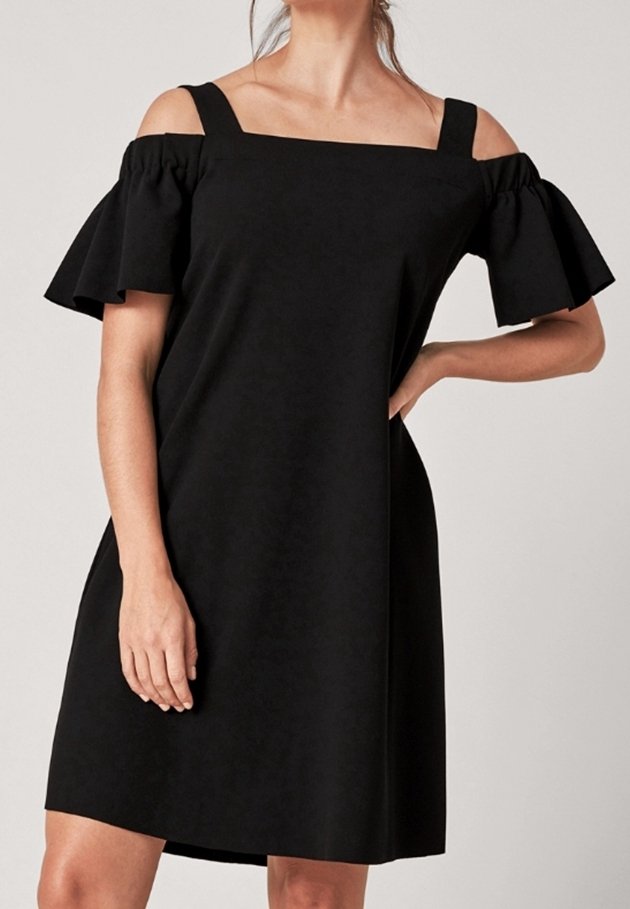 Femme portant une robe noire, longueur genou, épaules dénudées avec larges bretelles et manches courtes évasées, debout avec une main sur la hanche.