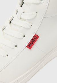 Witte sneaker van glad leer, met witte veters, een rode "HUGO" tag en een eenvoudige rubberen zool. Minimalistisch ontwerpdetail.