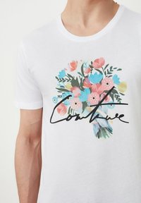 Valkoinen puuvillainen t-paita, jossa on kukkakuvio vaaleanpunaisilla, sinisillä ja vihreillä kukilla, sekä sana "Continue" mustalla käsinkirjoitetulla tekstillä.