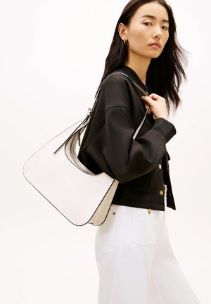 Donna con lunghi capelli neri porta una grande borsa bianca sulla spalla, indossa giacca nera e pantaloni bianchi su sfondo chiaro.