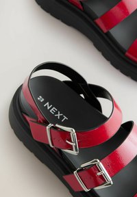 Sandales en cuir verni rouge avec deux sangles à boucle, semelle en caoutchouc noire et branding "NEXT" visible sur la semelle intérieure. Texture lisse dans l'ensemble.