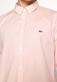 Camicia button-up rosa chiaro in cotone, con colletto a punta, bottoni bianchi e un piccolo logo di coccodrillo verde sul petto.