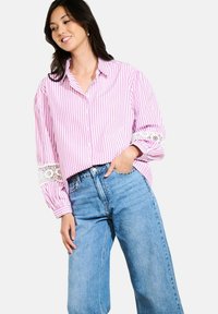 Woman wearing pink and white striped button-up shirt with lace detail on sleeves and high-waisted light blue wide-leg jeans, hand in pocket.