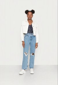 Casaco de ganga branco cortado sobre uma blusa floral azul-marinho, combinado com jeans retos claros rasgados e ténis brancos.