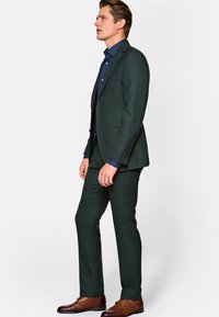 Ciemnozielony elegancki garnitur o slim fit, z teksturowanej tkaniny, zestawiony z niebieską koszulą. Brązowe buty typu double-monk dopełniają całość.