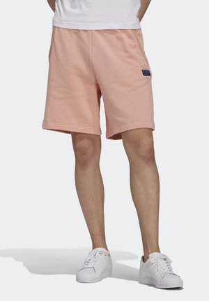 Mężczyzna w jasnoróżowych szortach Adidas z czarnym logo na prawej thigh i białych sneakersach, stojący na jednolitym tle.