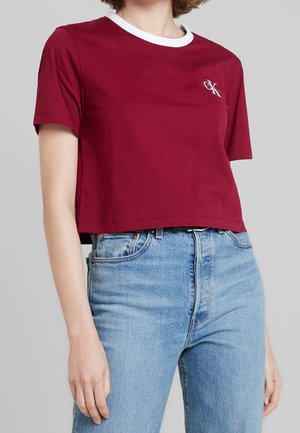 Frau trägt ein bordeauxfarbenes, kurzes T-Shirt mit weißem Kragen und kleinem CK-Logo, kombiniert mit hochtaillierten hellblauen Jeans.