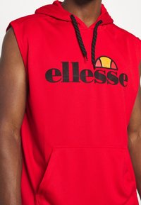 Red sleeveless hoodie with black branding "ellesse" on the front, featuring a multi-colored graphic above. Includes a drawstring hood.
