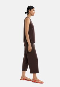 Brązowy bezrękawnik i szerokie spodenki sięgające do kostek, zestawione z czerwonymi sandałami na pasku. Gładki materiał, luźny krój i dyskretne rozcięcia po bokach.