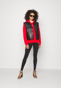 Zwarte leren vest over een rode hoodie, gecombineerd met zwarte legging met witte zijkanten en zwarte hakken. Accessoires zijn onder andere een paar zonnebrillen.