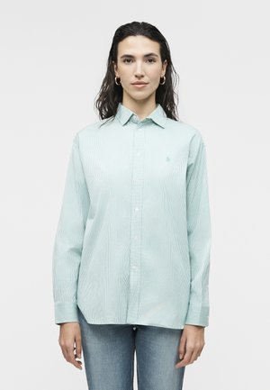 Vrouw met donker haar, gouden oorringen in de vorm van ringen, een lichtgroene gestreepte overhemd met knopen en een blauwe spijkerbroek, staand tegen een effen achtergrond.