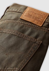 Rjavi džins hlače imajo usnjeno oznako z vtisnjenim logotipom. Tkanina ima teksturirano pletenje in kontrastne oranžne šive.