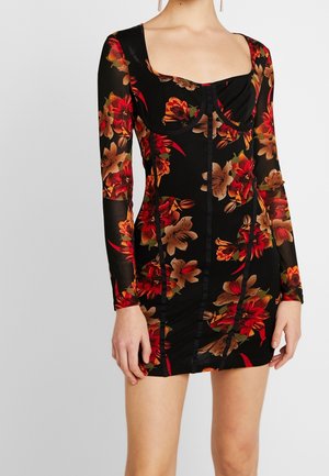 Kvinde iført en sort tætsiddende minikjole med lange ærmer med røde, orange og beige blomsterprint og en firkantet udskæring.