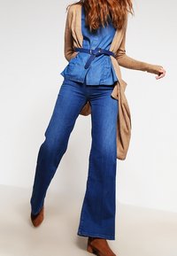 Jeansen är i denim och utsvängda, kombinerade med en blå peplumtop som är lagd under en lång beige cardigan. Outfitten har ett blått bälte som accent.