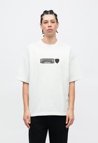 T-shirt bianca in cotone con vestibilità ampia, decorata con una grafica nera raffigurante un tram e un cuore sul davanti. Scollo rotondo. Maniche corte.