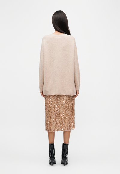 Μπεζ πλεκτό πουλόβερ με άνετη εφαρμογή, συνδυασμένο με φούστα μολύβι σε ροζ-χρυσό με παγιέτες. Μαύρα μποτάκια με λεπτή σόλα συμπληρώνουν την εμφάνιση.