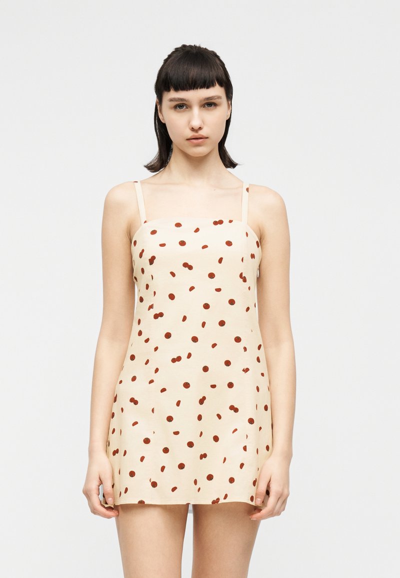 Νεαρή γυναίκα που φοράει μίνι αμάνικο φόρεμα σε εκρού χρώμα με λεπτές τιράντες και καφέ πουά, στέκεται μπροστά σε απλό φόντο.