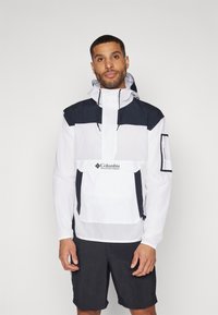 Hvid anorakjakke med en navy topsektion og en frontlomme, med justerbar hætte og elastiske manchetter. Bæret med mørke shorts.