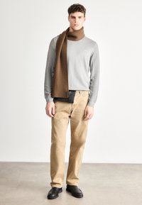 Bluză gri cu mâneci lungi și textură reiată, eșarfă tricotată maro, pantaloni bej cu croială dreaptă și pantofi slip-on negri. Design minimal cu accente.