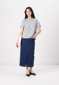 Un tricou gri cu mânecă scurtă, asociat cu o fustă din denim albastru închis, cu un slit diagonal în față și un tiv curat. Modelul ține o geantă albă.