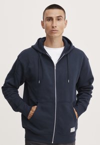 Marineblå zip-opp hettegenser med hette, frontlommer og snorer. Laget av myk stoff, med en jevn tekstur og ribbestrikkede mansjetter.