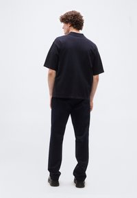 Άνδρας με σγουρά μαλλιά στέκεται με πλάτη, φοράει ένα μαύρο κοντομάνικο polo πουκάμισο, σκούρο παντελόνι και μαύρα παπούτσια σε φωτεινό φόντο.