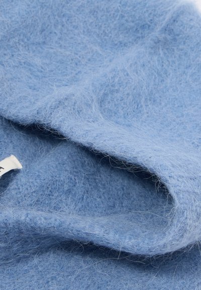 Tecido de lã azul claro com uma textura suave e fofa, mostrando fibras finas e dobras suaves. Uma etiqueta é visível em uma das bordas.