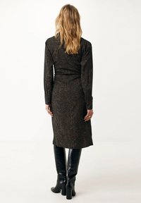 Femme avec des cheveux blonds ondulés portant une robe à manches longues, jusqu'au genou, à motifs et des bottes noires à talons hauts arrivant au genou, de dos.