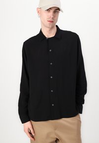 Schwarzes Langarmshirt aus weichem Stoff, mit einer Knopfleiste, klassischem Kragen und lockerer Passform. Getragen mit beigen Hosen.