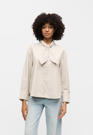 Γυναίκα με σγουρά μαλλιά που φοράει μπεζ πουκάμισο με κουμπιά και λεπτομέρεια με γραβάτα στο λαιμό και ανοιχτό μπλε τζιν, που στέκεται μπροστά σε απλό λευκό φόντο.