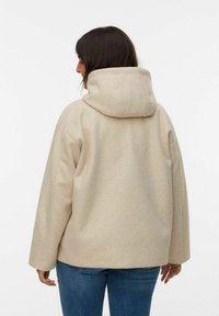 Beige hættejakke med en blød tekstur. Har en afslappet facon og sænkede skuldre, parret med tætsiddende blå jeans.