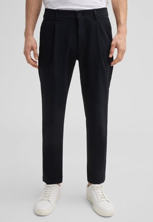 Schwarze Hosen aus strukturiertem Stoff, mit Falten, Knopfverschluss und elastischem Bund. Kombiniert mit weißen Sneakern.