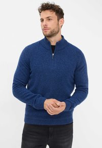 Maglione blu lavorato a maglia con collo alla zip e polsini a coste. Tessuto morbido, di media spessore. Indossato sopra una camicia nera.