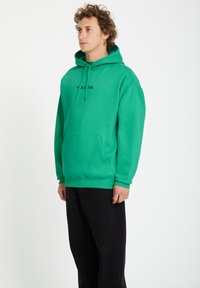 Groene hoodie van zachte stof, met een kangoeroezak en een capuchon met trekkoord. Aan de voorkant staat 'VOLCOM' in zwarte tekst. Draagt met zwarte broek.