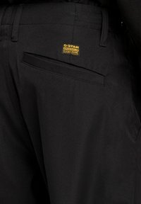 Svarta byxor med en slät textur, med en G-Star-etikett i guld. Inkluderar en bakfick och en skräddarsydd design.