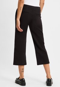 Black cropped trousers with a smooth texture, featuring a fitted waistband, two back pockets, and paired with chunky black shoes.